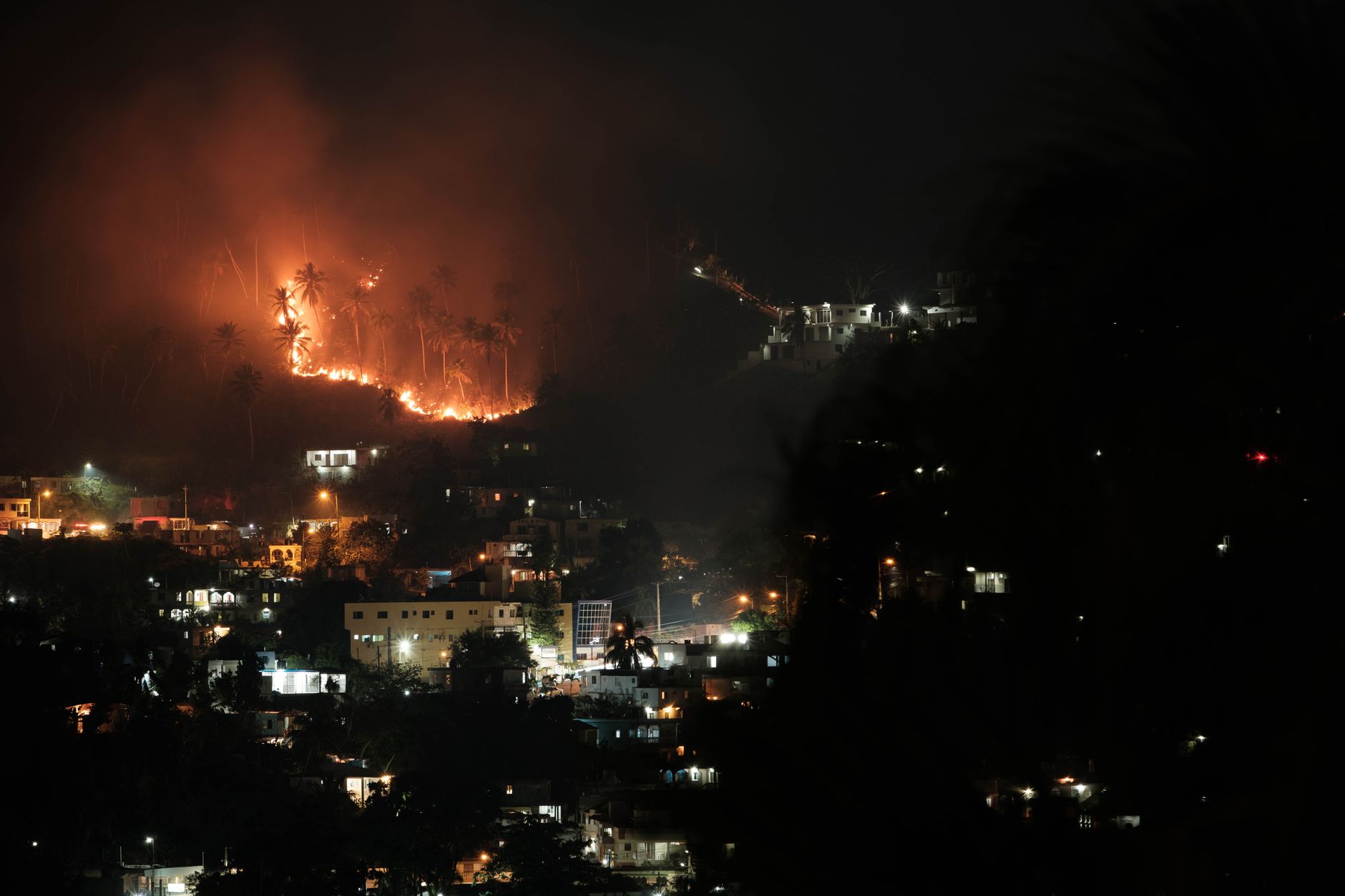 Aerial view of a forest fire at night near a town, with bright flames lighting up the sky and smoke billowing upwards.